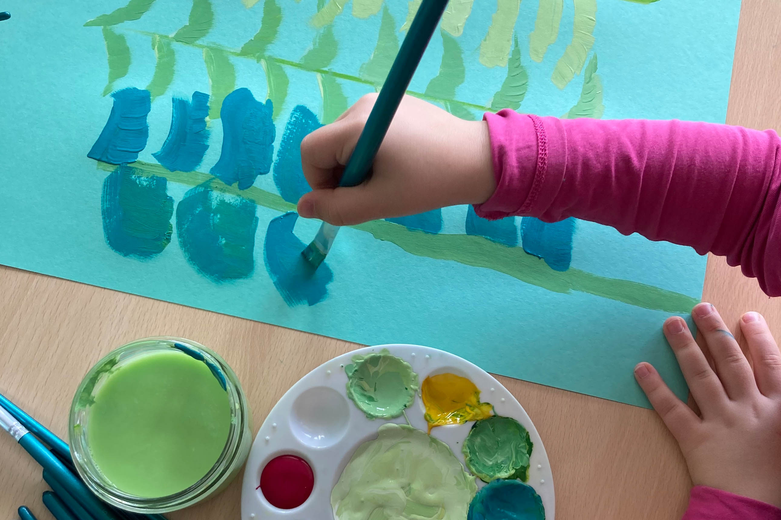 Gros plan : main d'enfant en train de peindre des sapins sur un papier bleu
